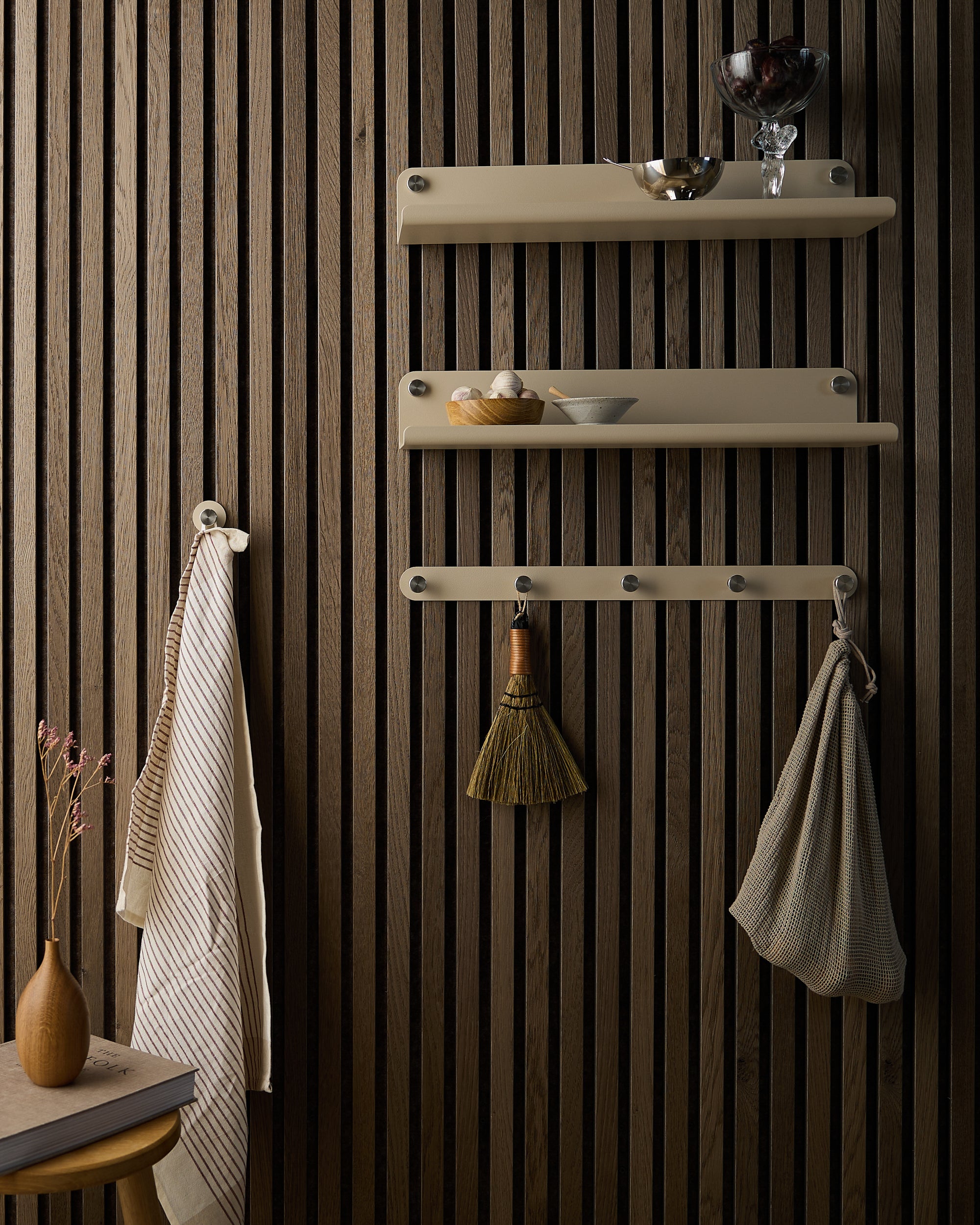Three beige wall shelves hold dishes and cups on a dark wood slat wall. Below, hooks display a broom and towels. A small wooden table with a vase and dried flowers sits nearby.