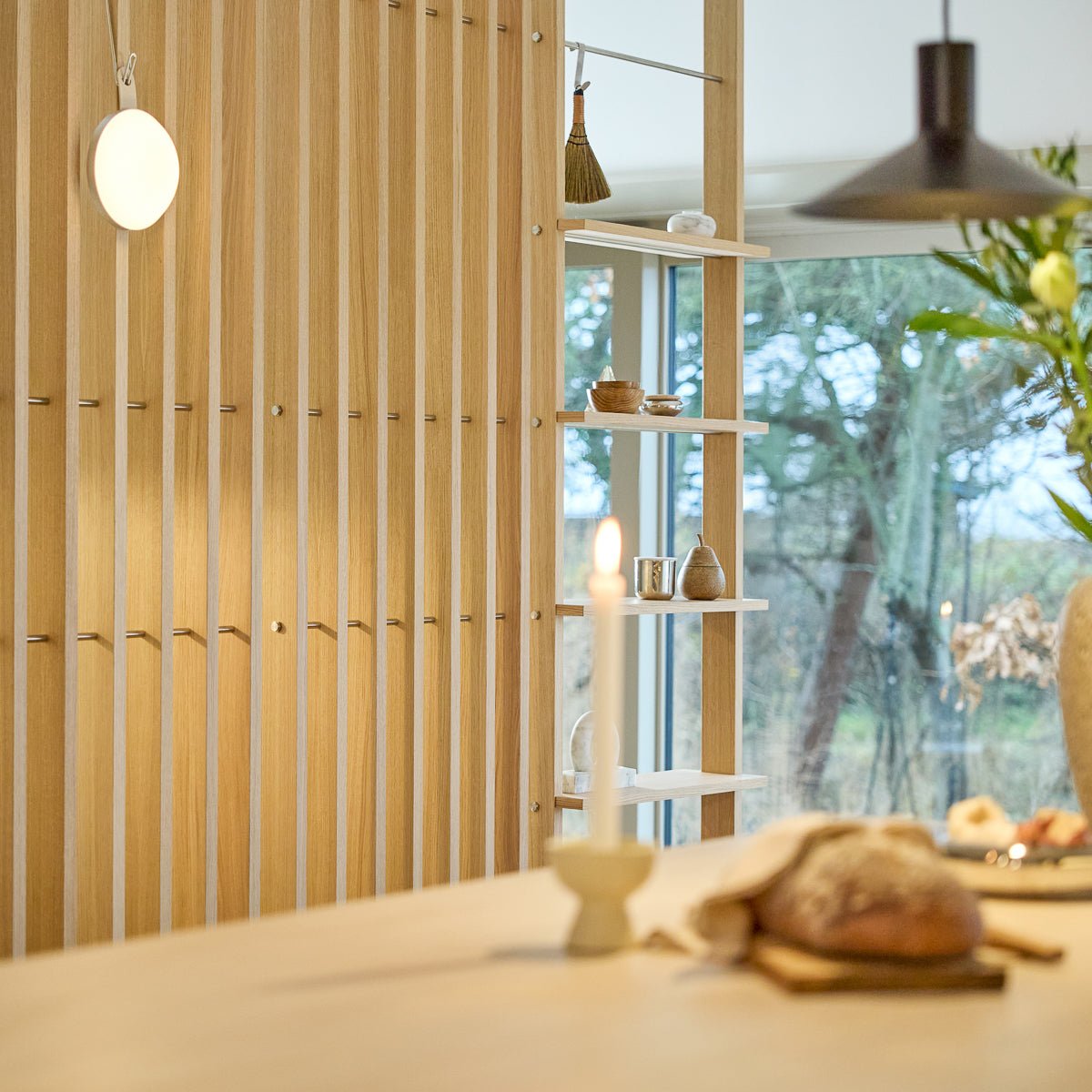 A lit candle and bread sit on a wooden table in a bright room with vertical wood slats, open shelving, and a large window showing trees outside. Scandinavian minimalist design.