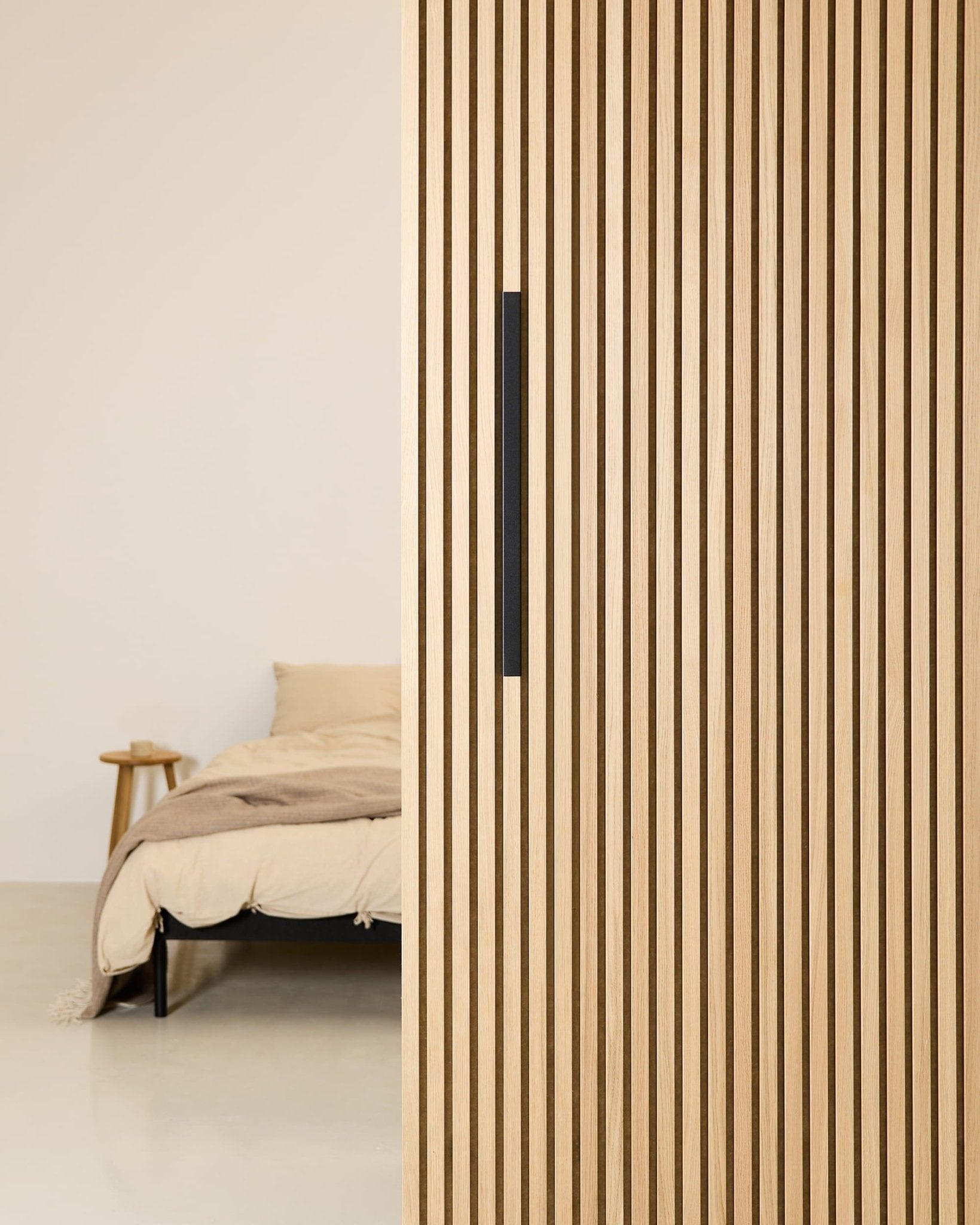 Minimalist bedroom with a beige bed, partially screened by a 60 cm WoodUpp EMBRACE black aluminum room divider featuring a black handle.