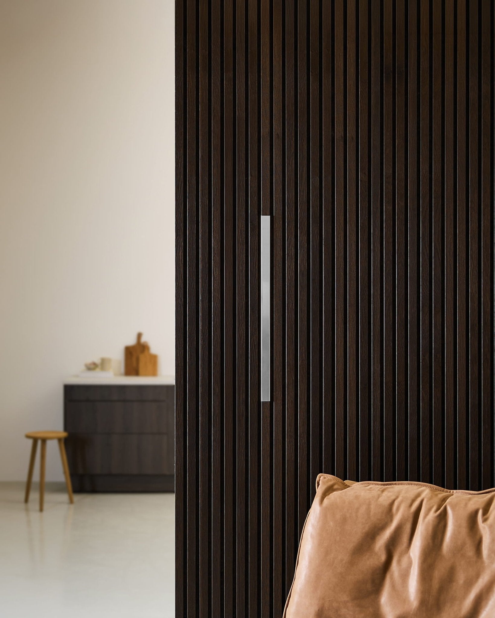 Modern Nordic space featuring WoodUpp Liva polished steel acoustic panels, a dark wood slatted wall, tan leather chair, and stool by a kitchen cabinet.