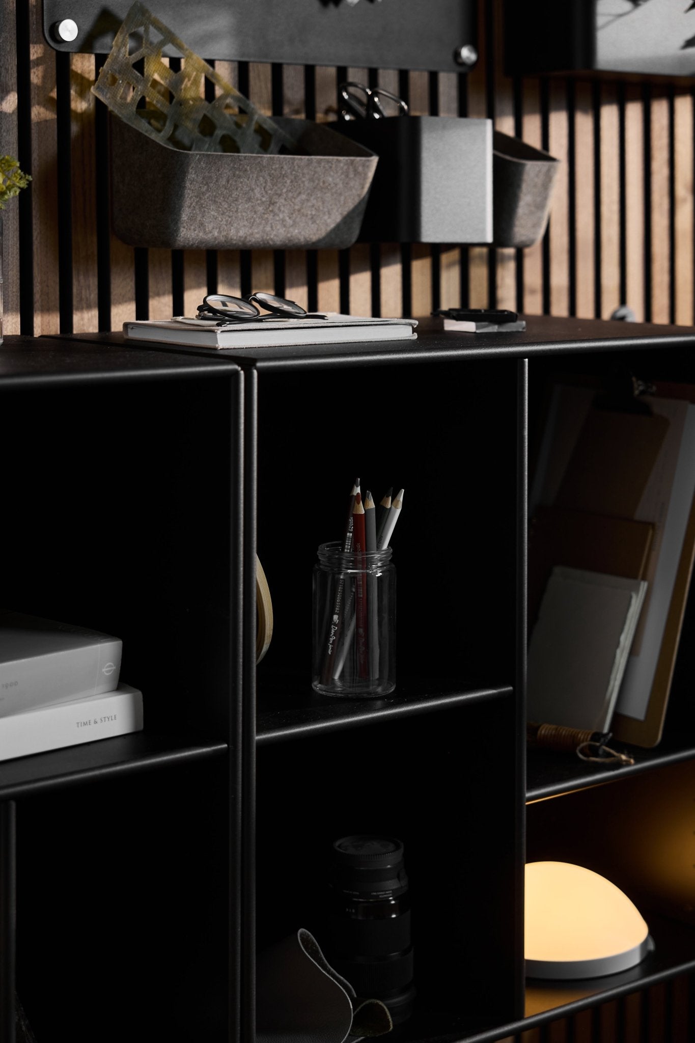 The Arka Open Cabinet in black by WoodUpp displays pencils, notebooks, books, a camera lens, a round lamp; organizers are mounted above.