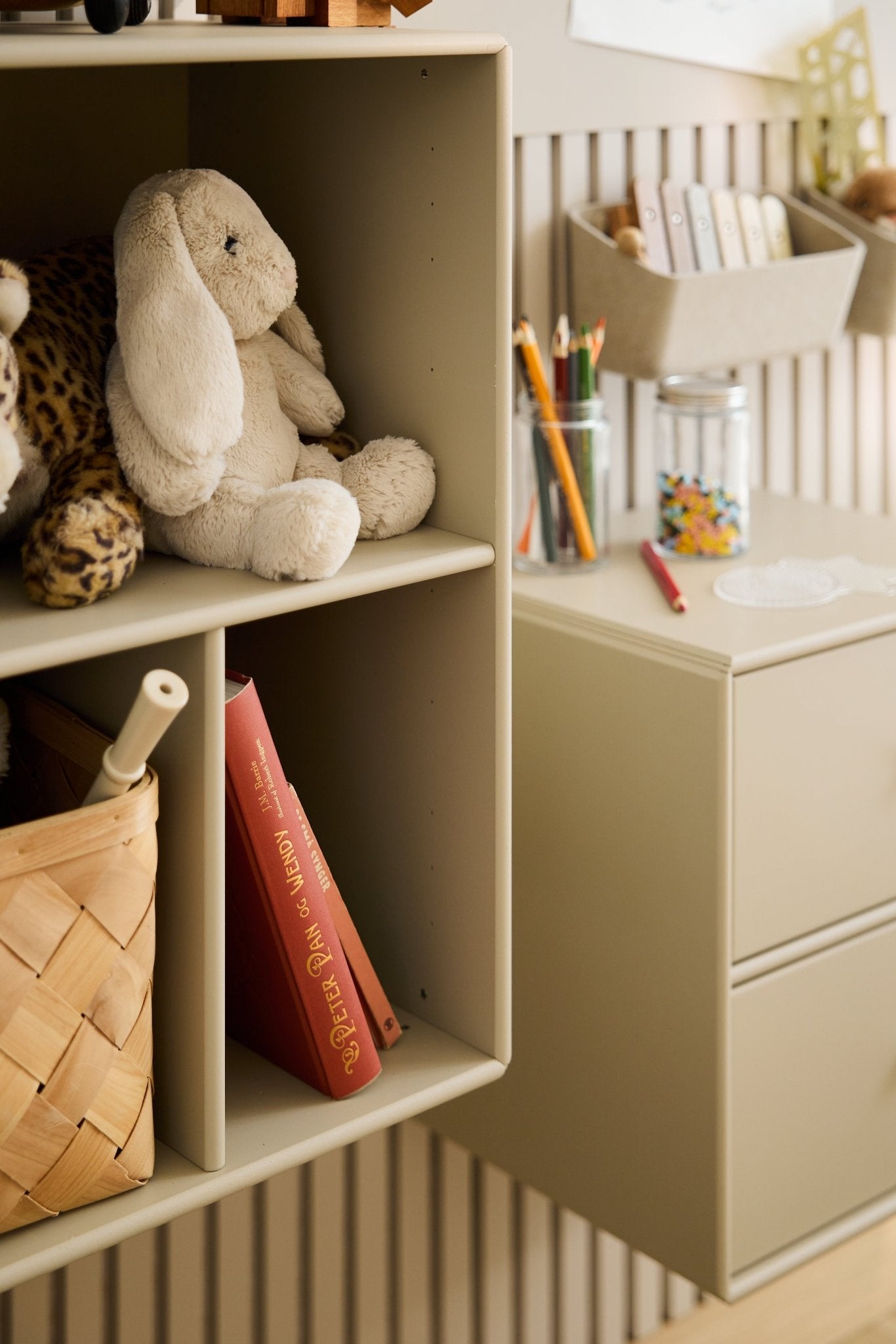 Stuffed animals, art, and a red book on a WoodUpp Arka open acoustic cabinet in beige, beside a wicker basket and floating drawer with beads.