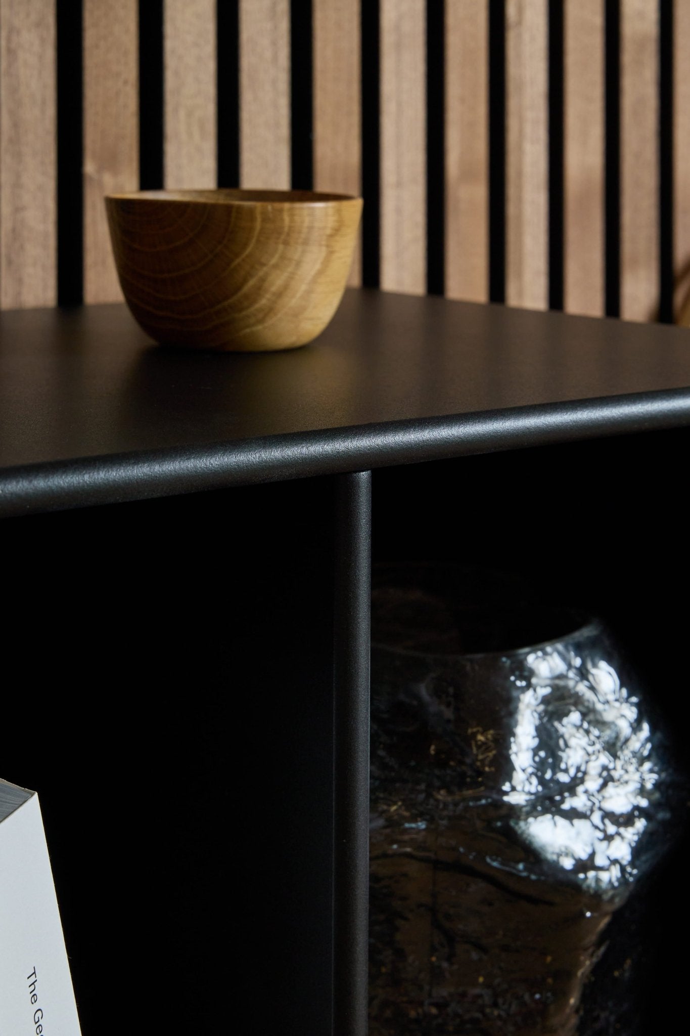 A wooden bowl on a black surface in front of WoodUpp Arka Bookcase / Black, with a glass object and book underneath, highlighting Nordic acoustic design.