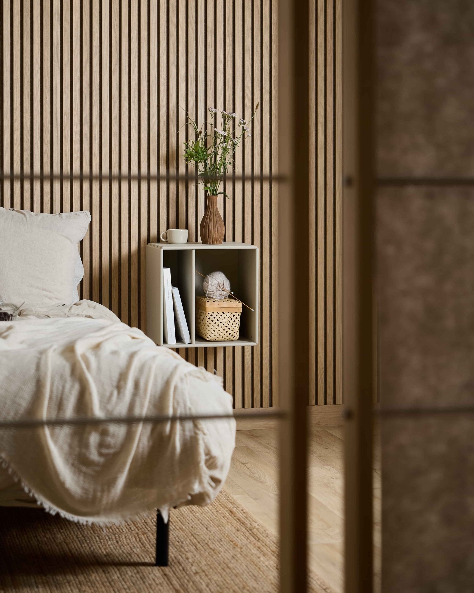 Minimalist bedroom with WoodUpp Arka Bookcase in beige, books, basket, and flower vase placed against a Nordic Akupanel acoustic slatwall panel.