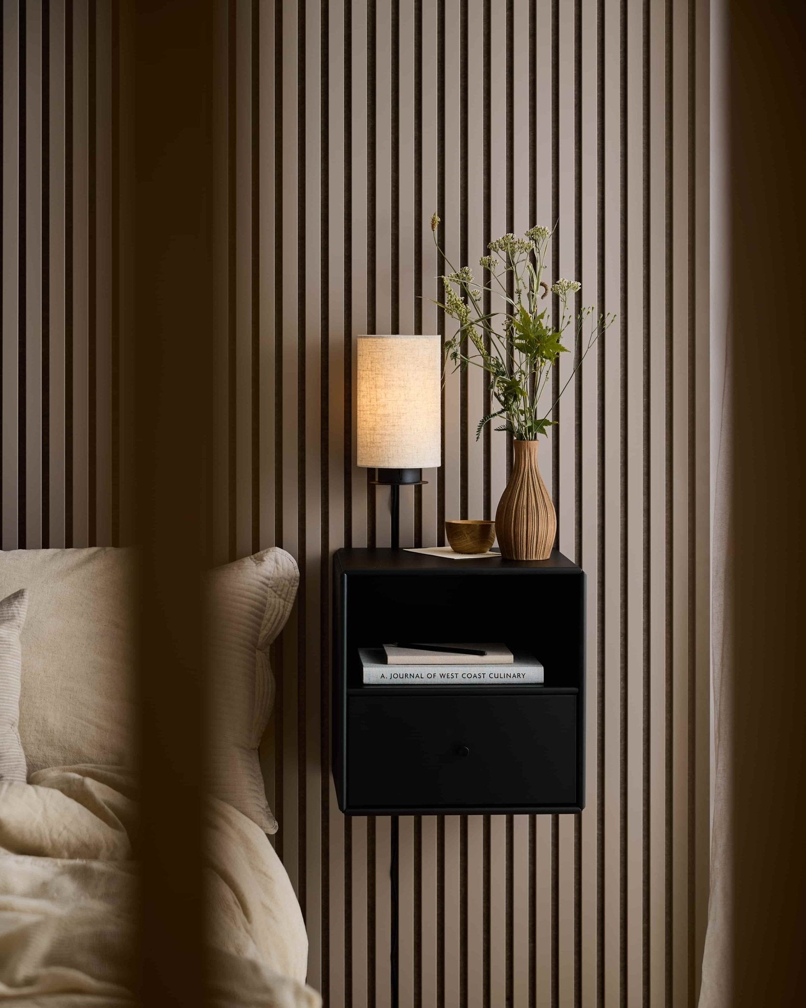 Bedside table with lamp, vase, cup, and book next to a bed and WoodUpp Akupanel acoustic panel in Dark Cashmere for sound absorption.