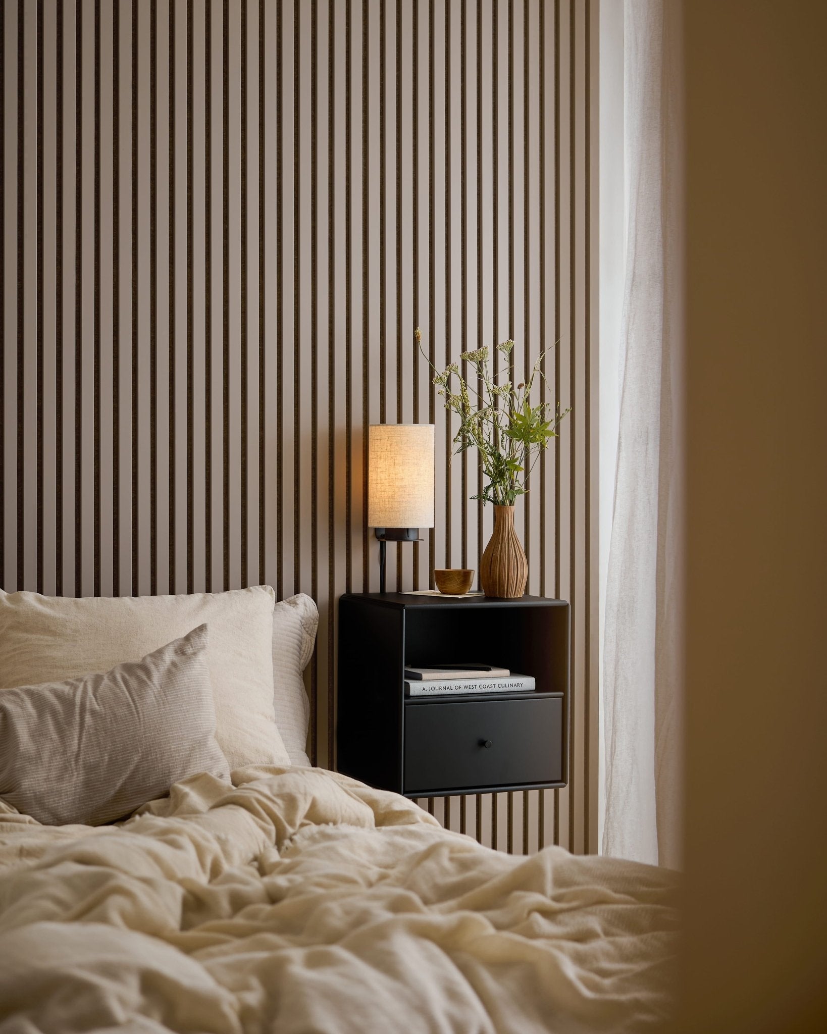 Bed with cream bedding, books, and lamp on black nightstand beside a WoodUpp Akupanel Dark Cashmere acoustic panel for style and sound dampening.