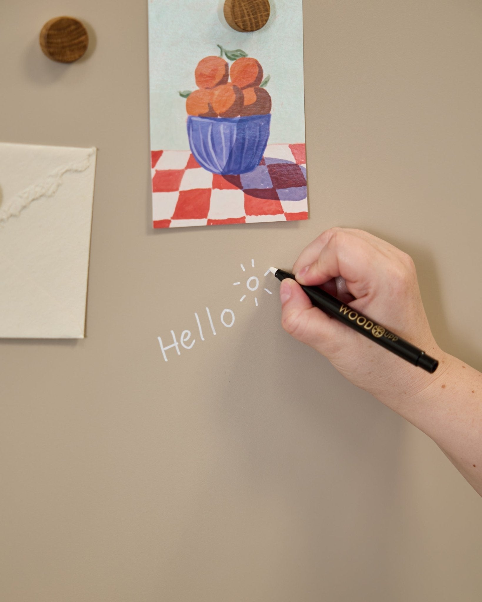 Hand writing hello with a white marker on a beige acoustic wall panel with art held by round wooden magnets