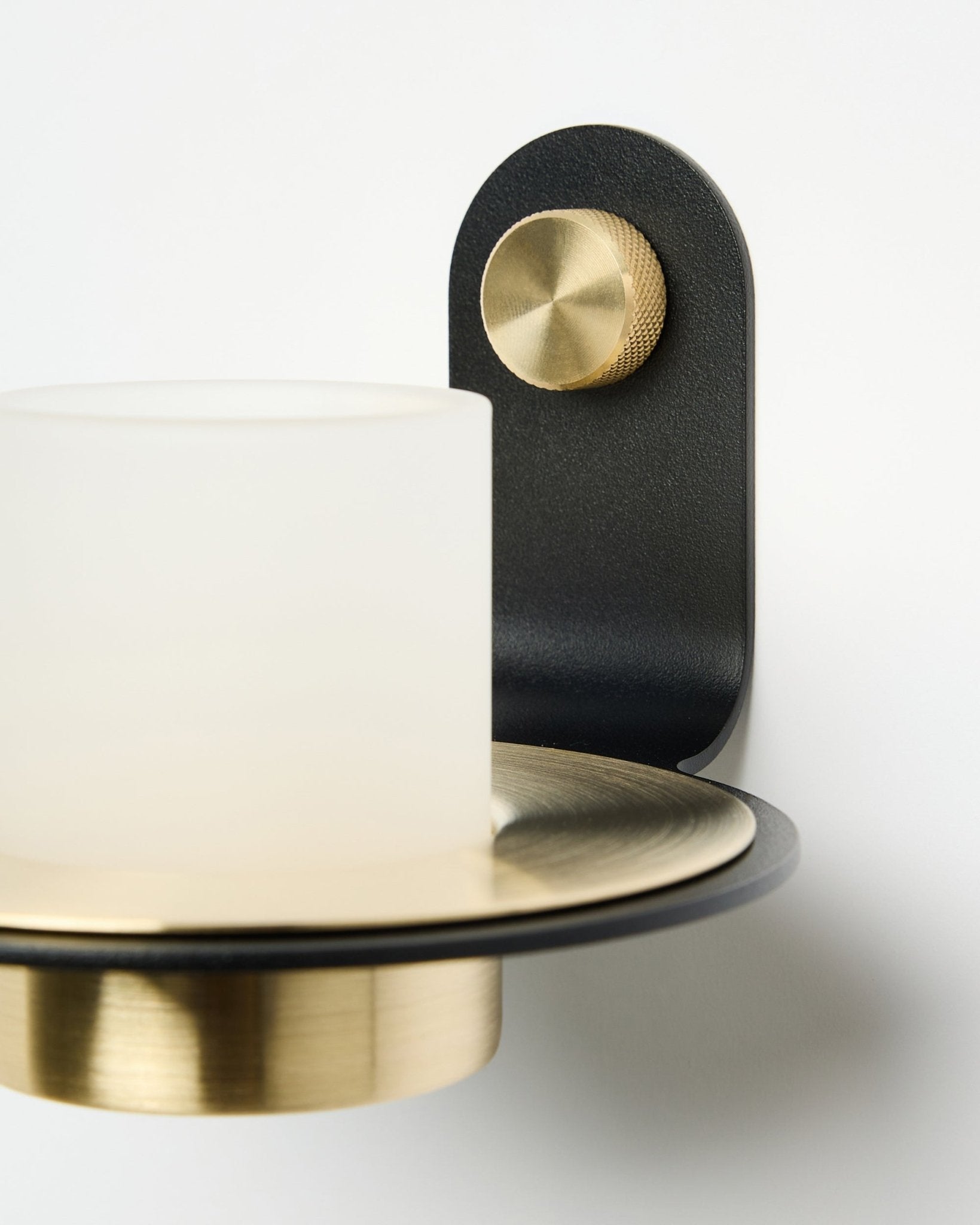 Black wall-mounted candle holder with brass knob and white glass shade on plain white background