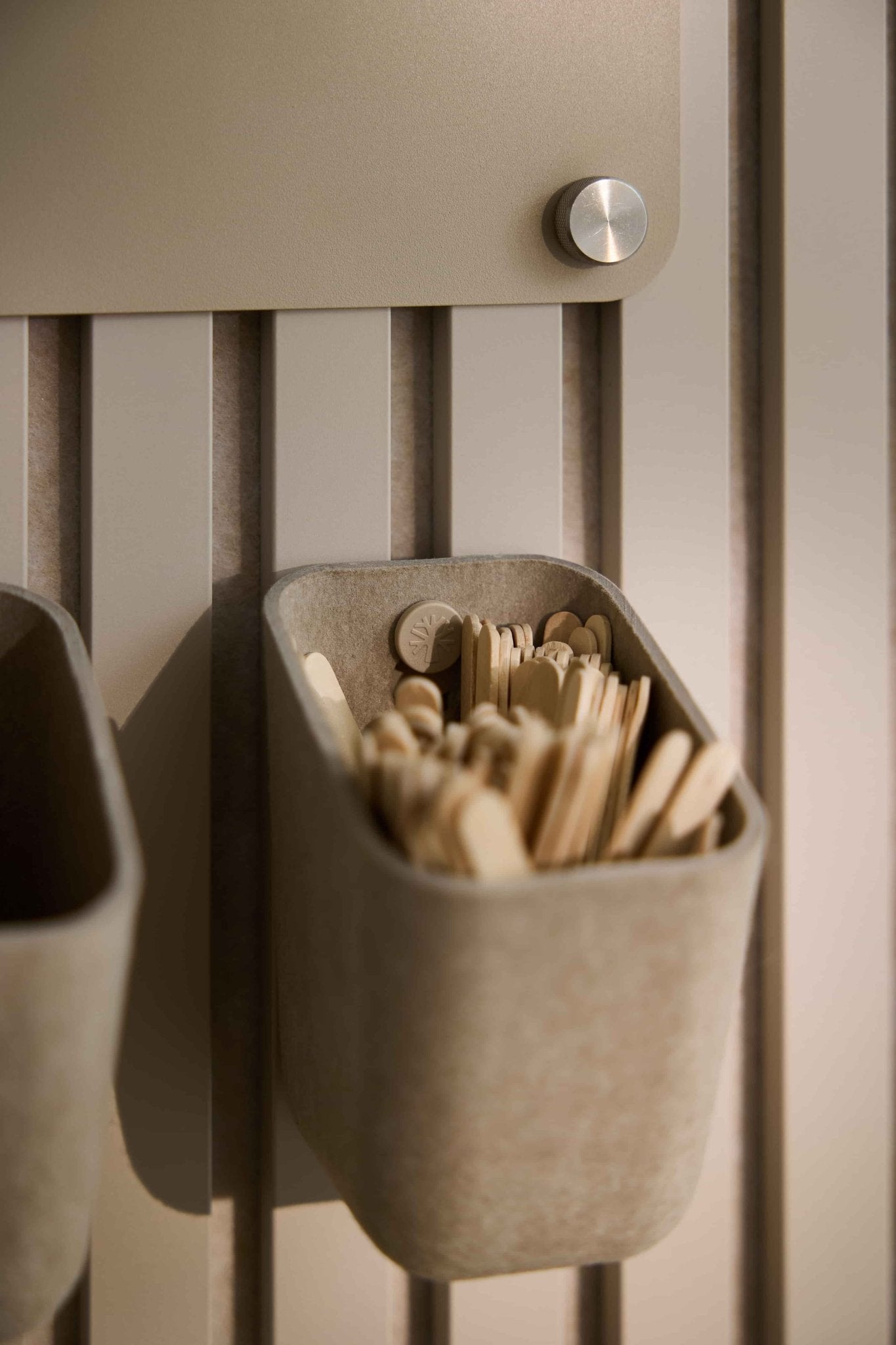 WoodUpp Dupps | Light organizer in beige holds stir sticks and spoons, mounted on a Nordic Akupanel acoustic panel with a metal knob above for access.
