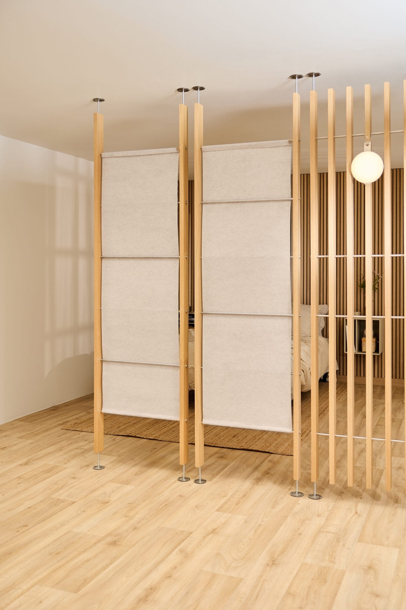 Modern room with light wood floors, a light beige WoodUpp EMBRACE felt sheet room divider, bed, and round wall lamp visible in the background.
