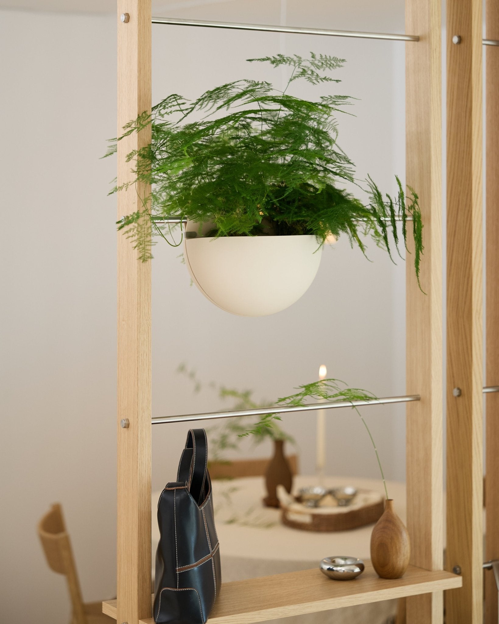 A green fern in a light beige WoodUpp Gro Soft Pocket BM2 planter rests on a wooden shelf above a black bag, vase, and set dining table.