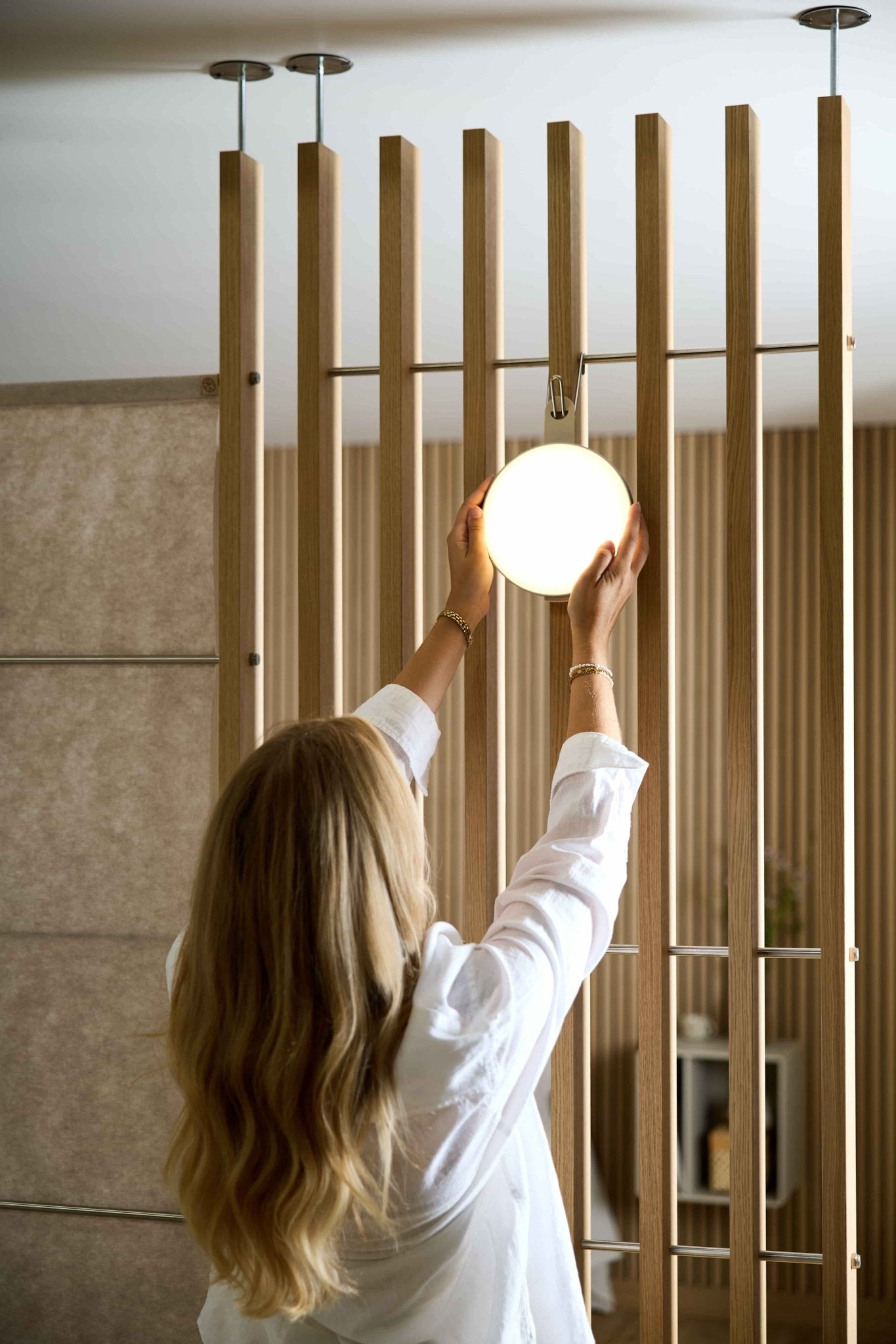 A woman with long blonde hair adjusts the Vala Portable LED Lamp by WoodUpp on an EMBRACE wooden slatted room divider in a modern interior.