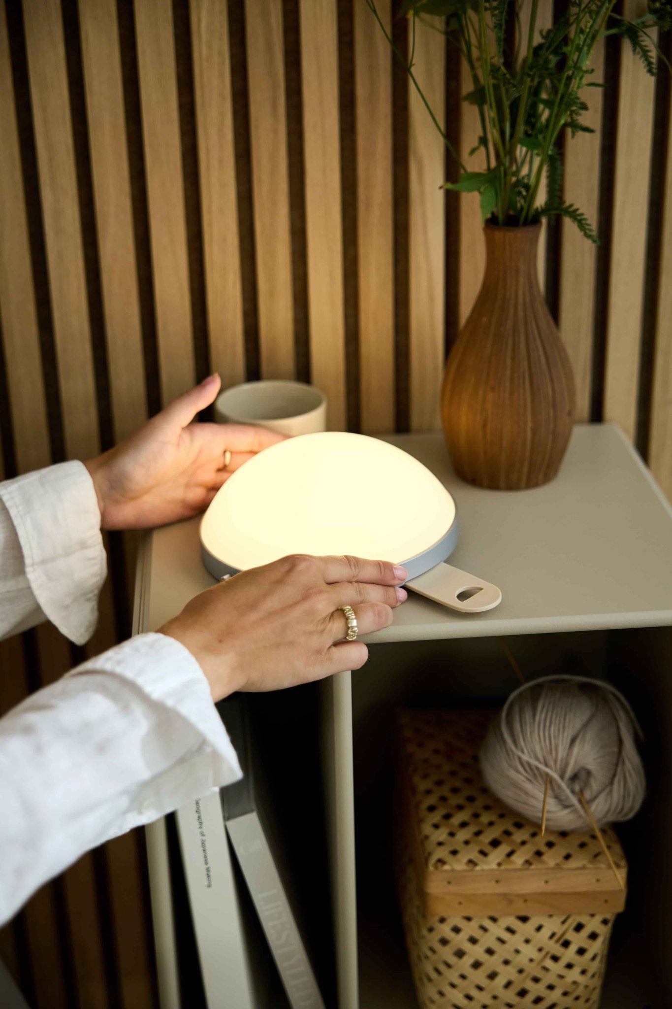 A person switches on the Vala | Portable LED Lamp by WoodUpp on a side table with a vase, cup, basket, and yarn ball next to an Akupanel acoustic panel.