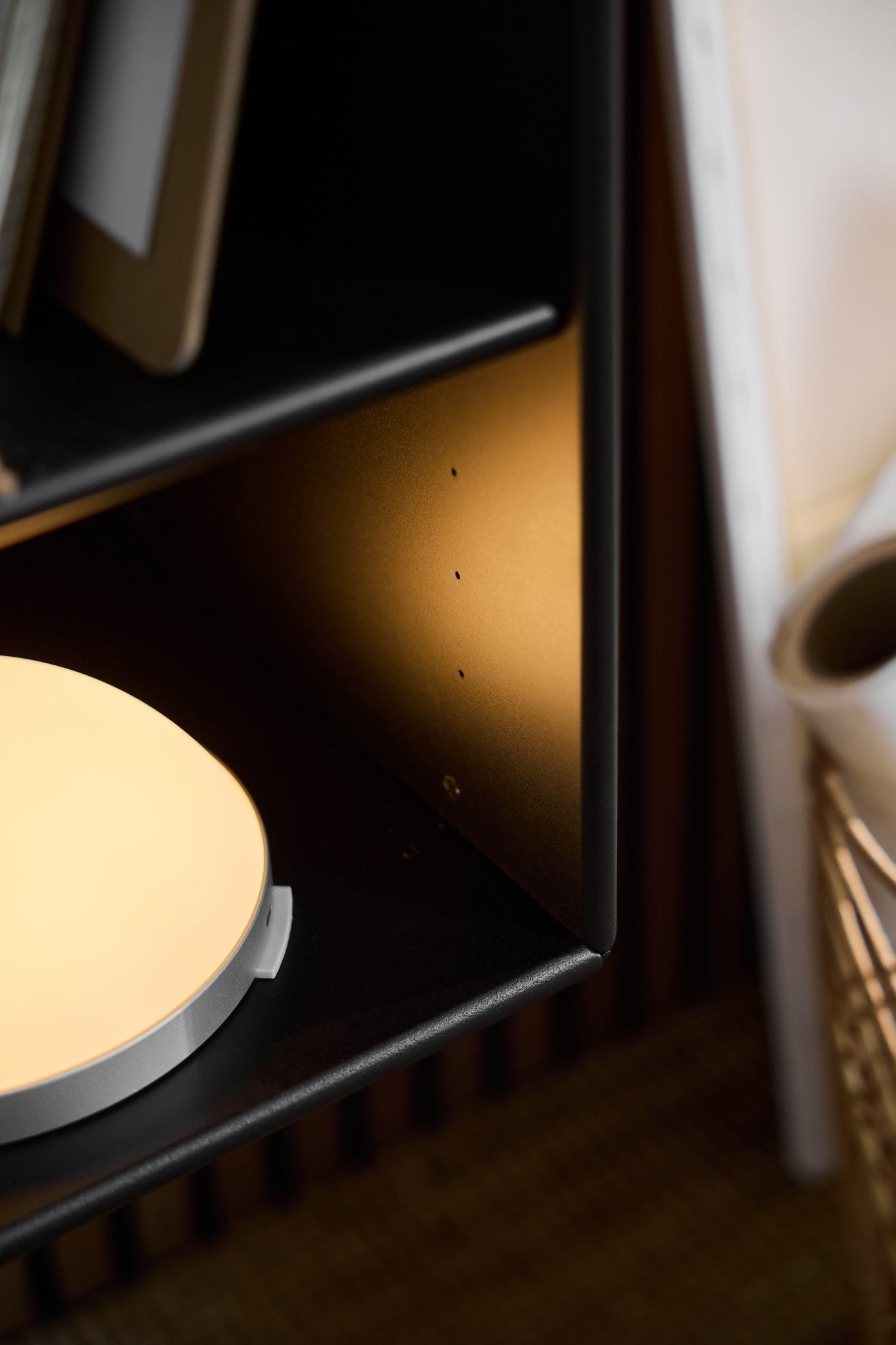 Close-up of the Vala portable LED lamp by WoodUpp on a black shelf, with blurred rolled paper and wire basket in the background.