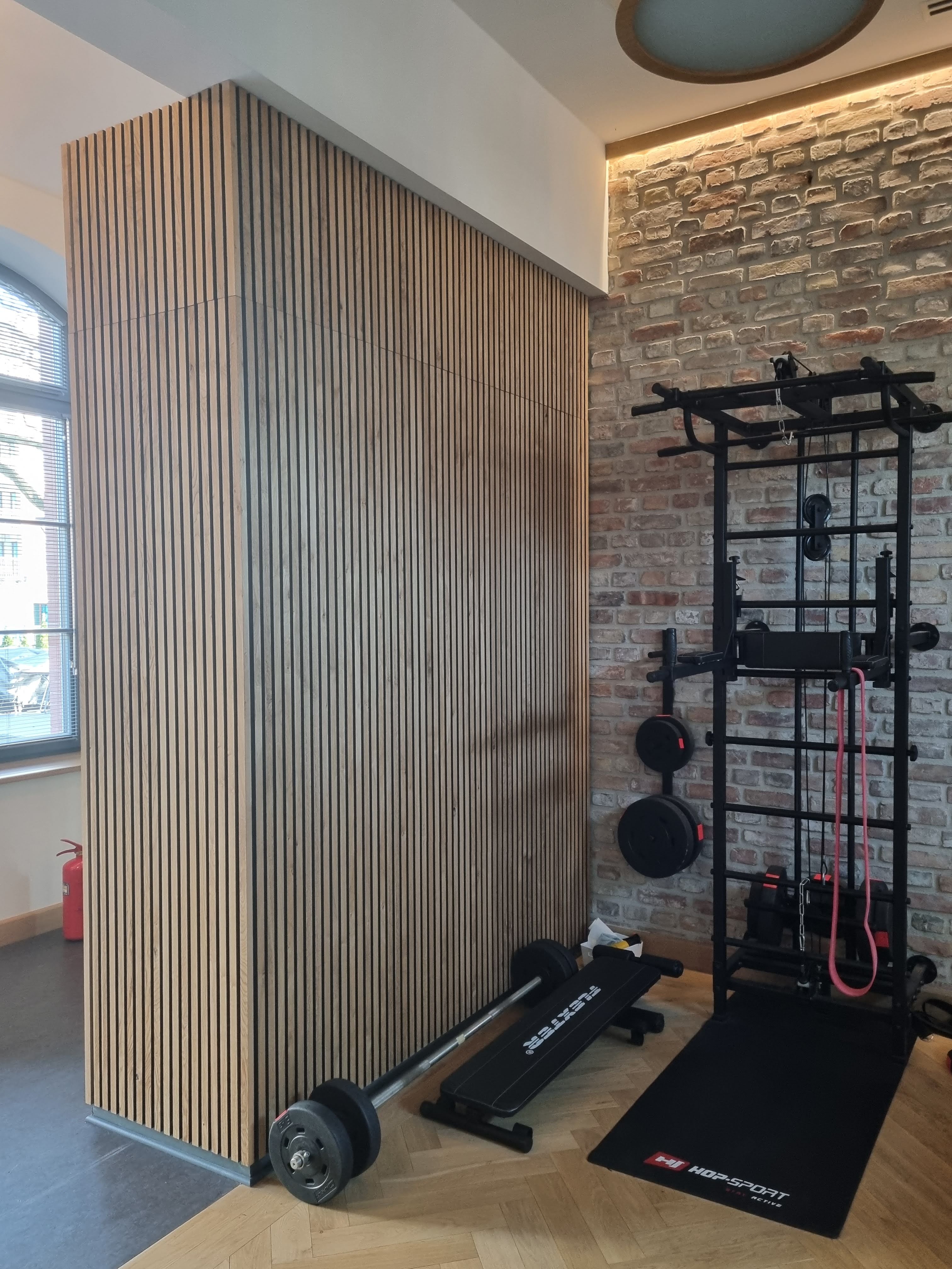 Akupanel acoustic slat panels covering a corner column in a gym interior with exposed brick wall and workout equipment.