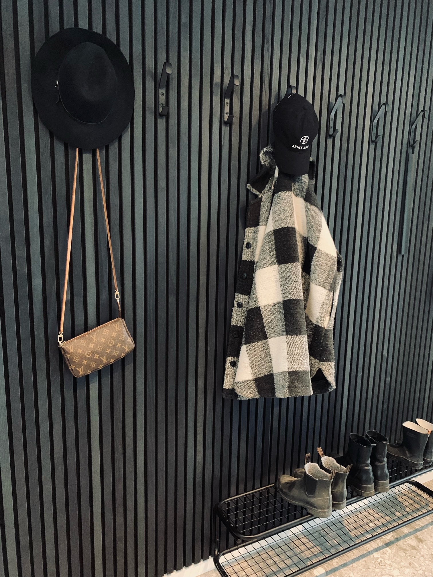 A black hat, plaid coat, cap, and brown purse hang on hooks with boots and shoes below, set against a dark Nordic acoustic slatwall panel.