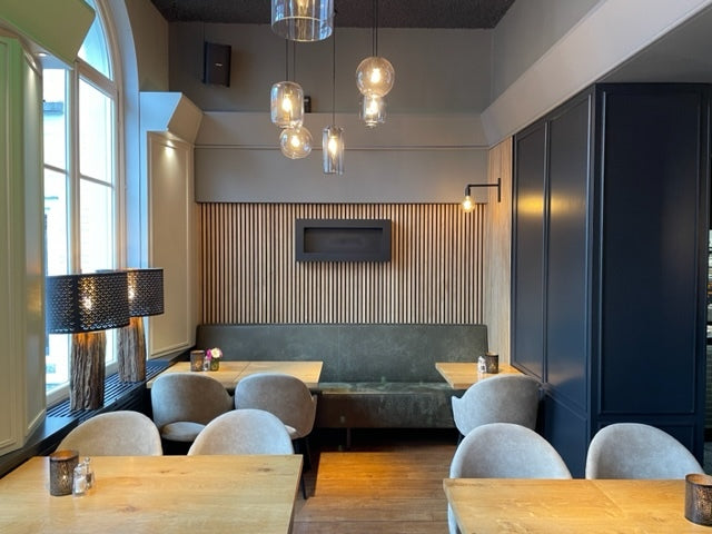 Modern cafe with wooden tables, beige chairs, pendant lights, and an Akupanel acoustic wood slatwall. Large windows provide ample natural light.