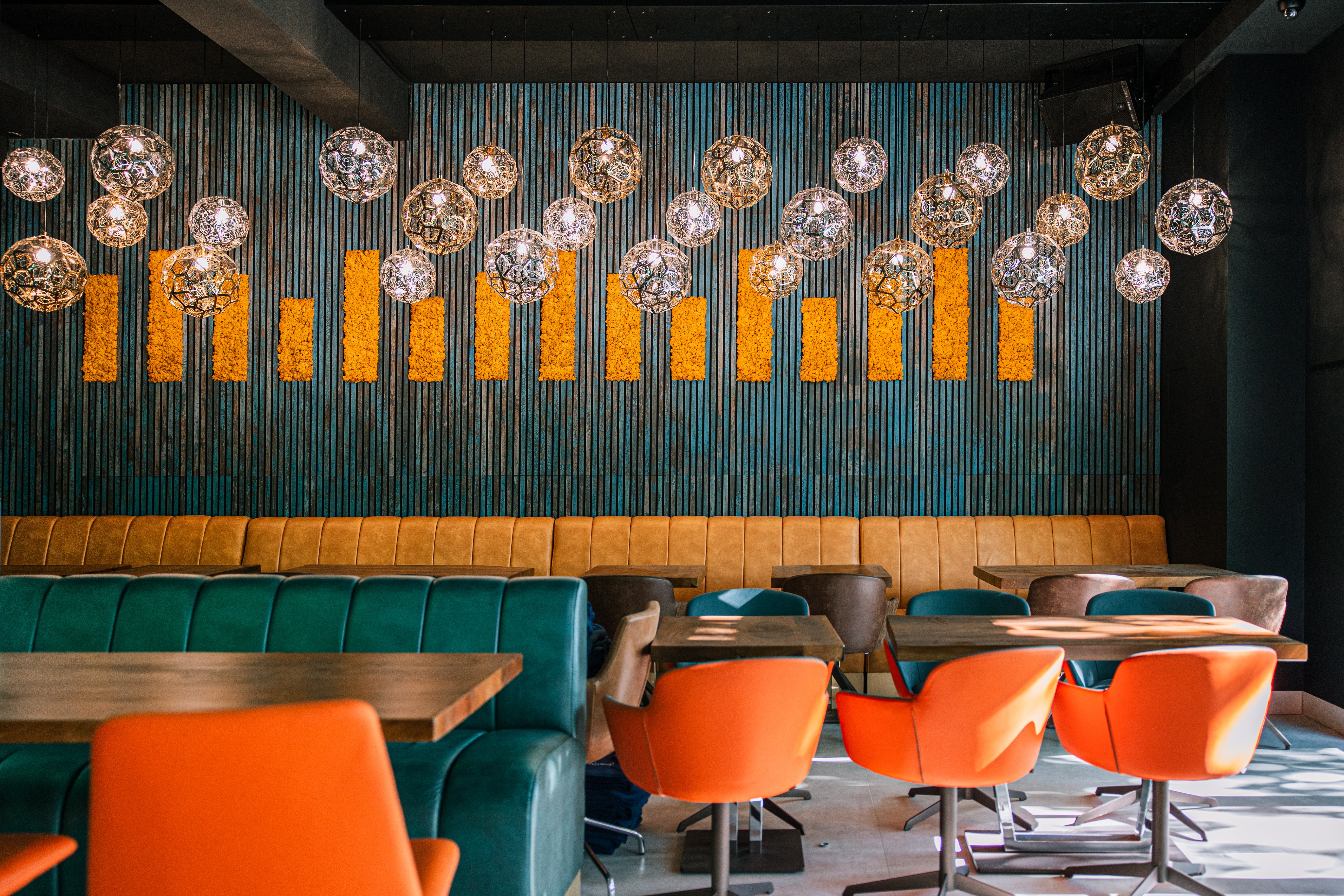A modern restaurant with teal booths, orange chairs, wood tables, and spherical pendant lights by a textured acoustic panel wall with yellow vertical accents.
