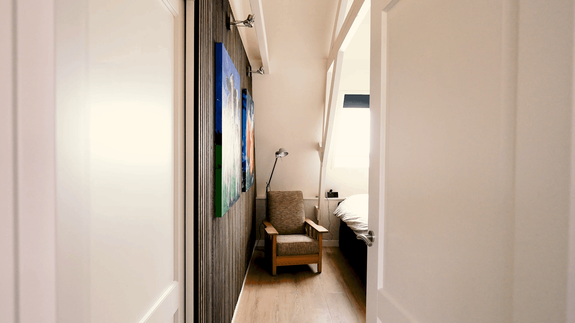 Akupanel acoustic slat wall panels, dark wood on black backing, beside armchair and paintings in a narrow bedroom.
