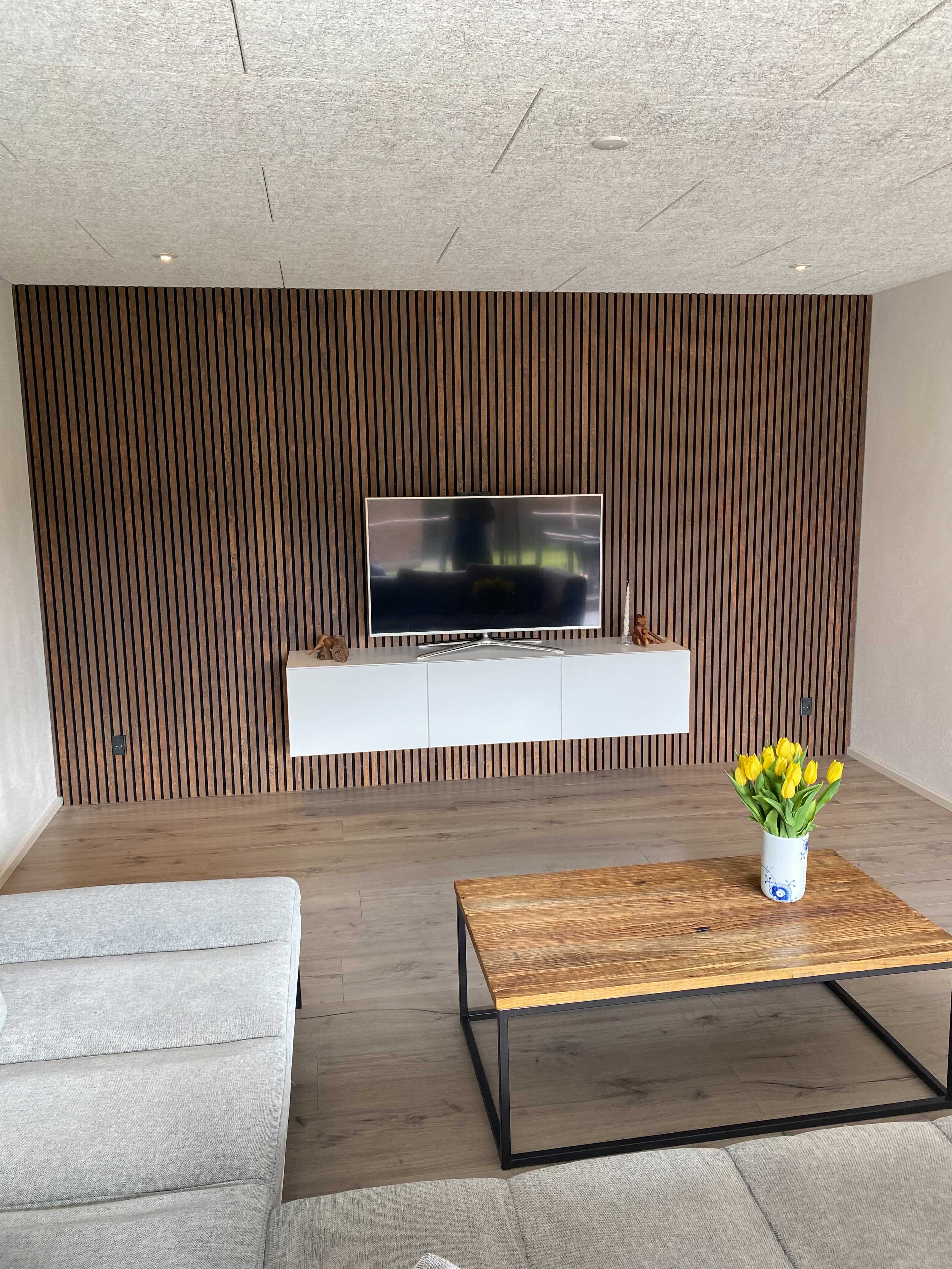 Modern living room featuring a TV on a Nordic-inspired acoustic wood slat panel, floating white console, gray sofa, wood coffee table, and yellow tulips.
