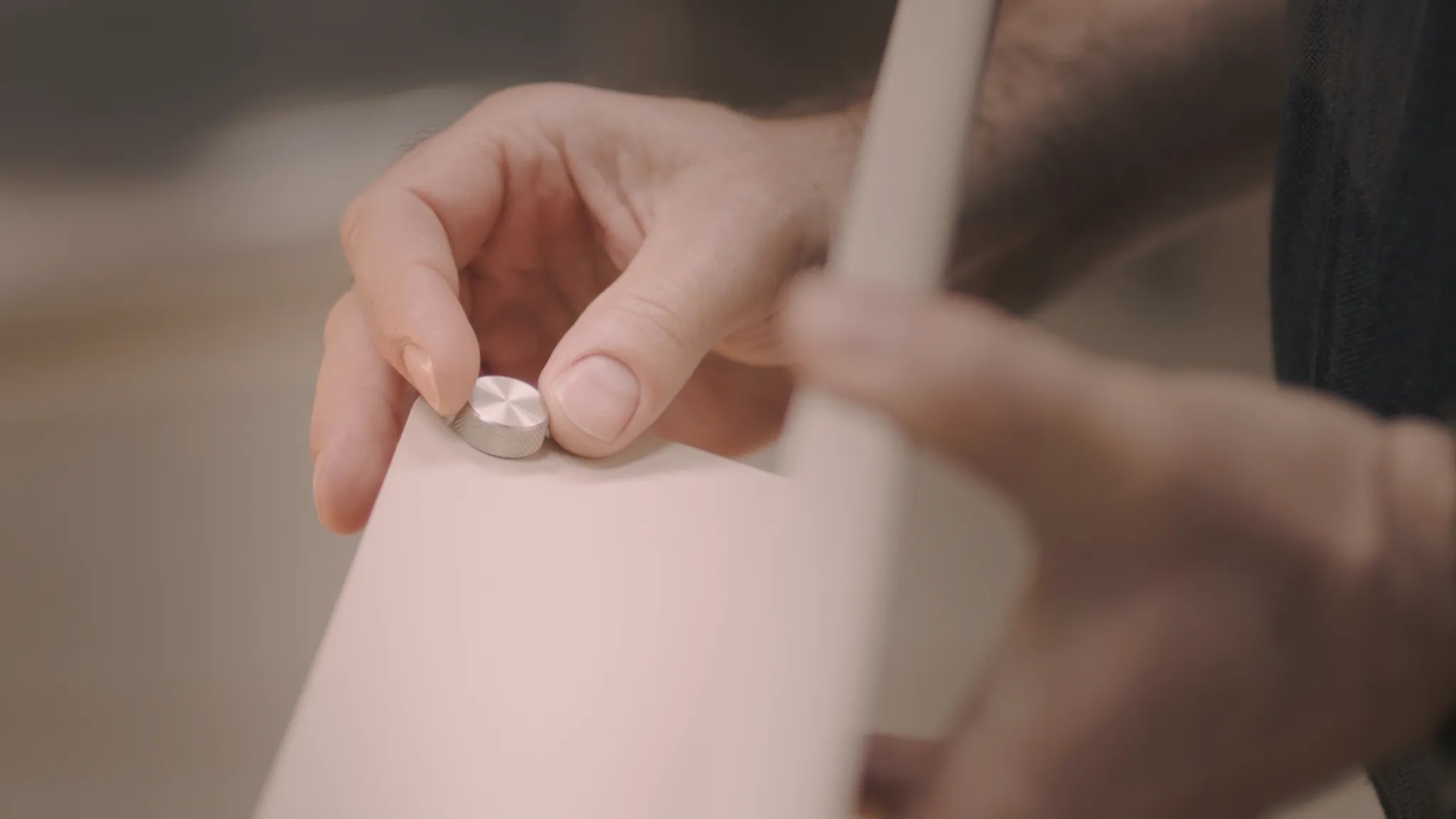 Round brushed metal mounting cover being fitted by hands onto a pale panel, blurred background