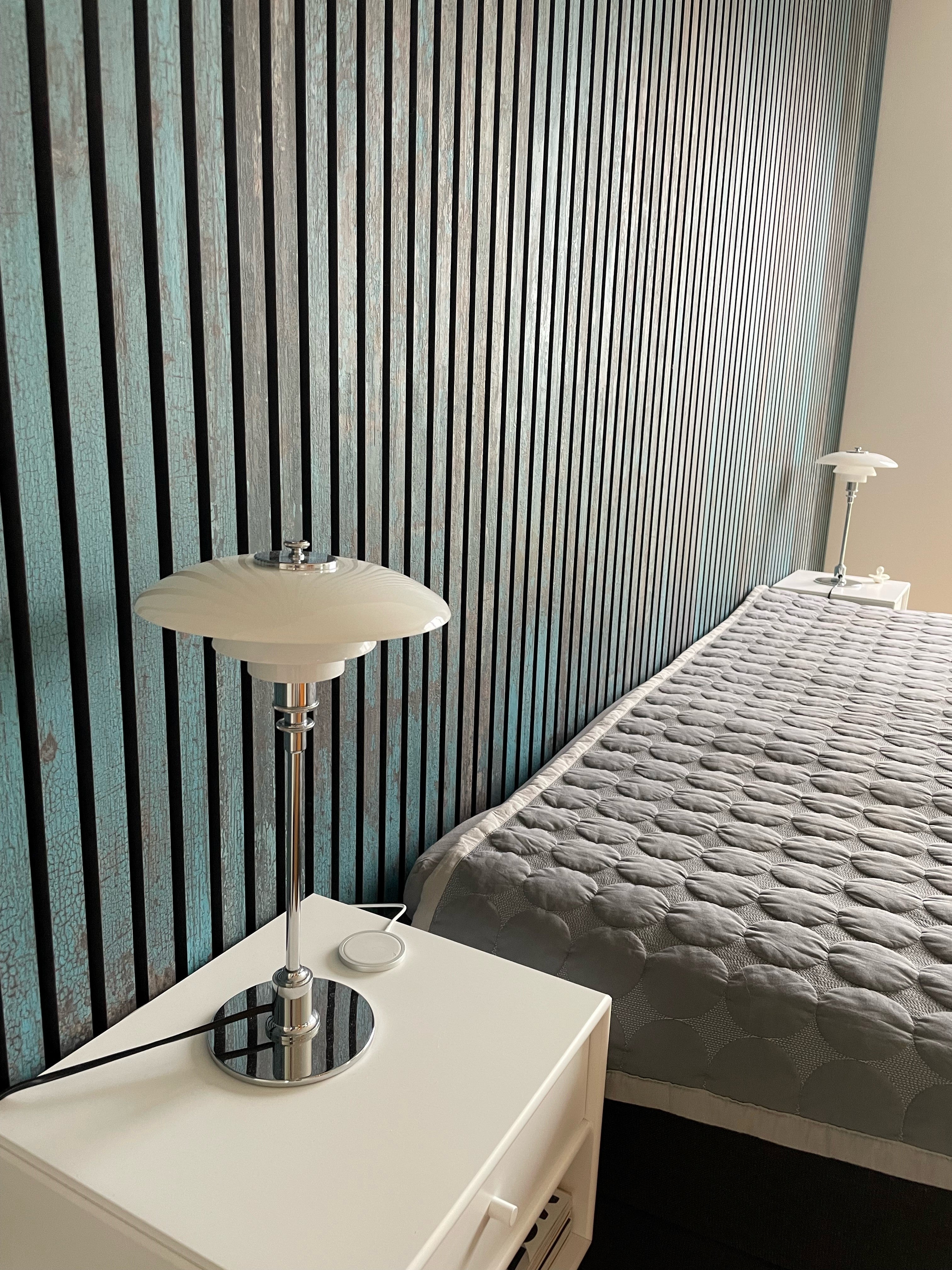 A modern bedroom with a gray quilted bed, white nightstand, and chrome lamp, featuring a blue-and-black striped Akupanel acoustic wall panel accent.