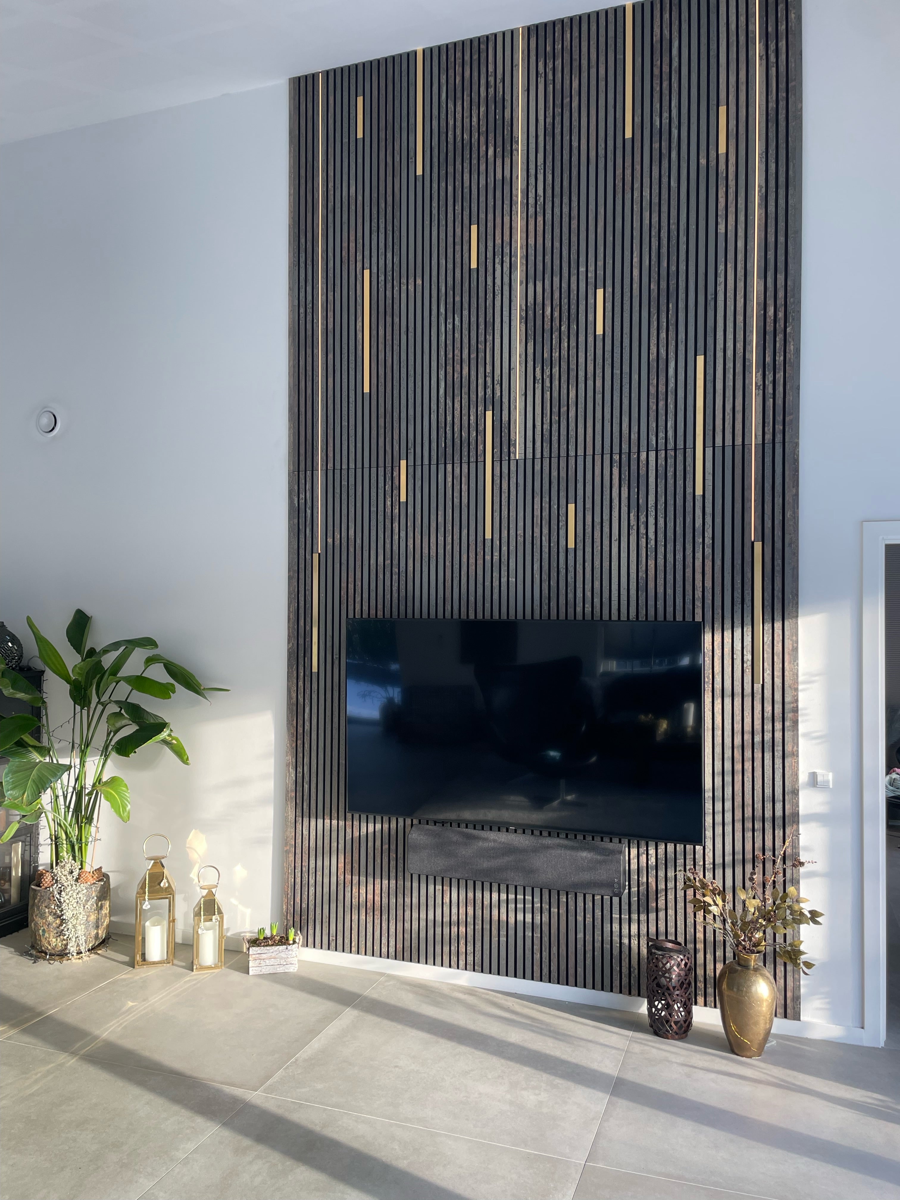 A modern living room with a wall-mounted TV on a dark acoustic wood panel, gold accents, plants, lanterns, and decorative vases.