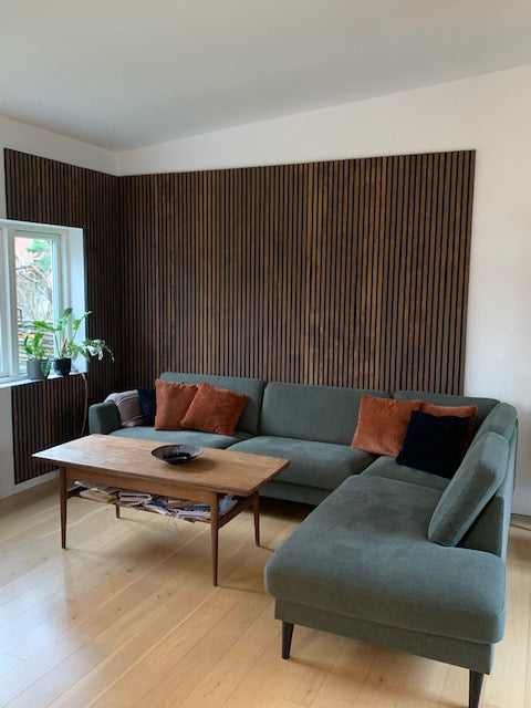 Modern living room featuring a green sectional sofa, cozy pillows, wooden coffee table, houseplants, and an acoustic slatwall panel accent.