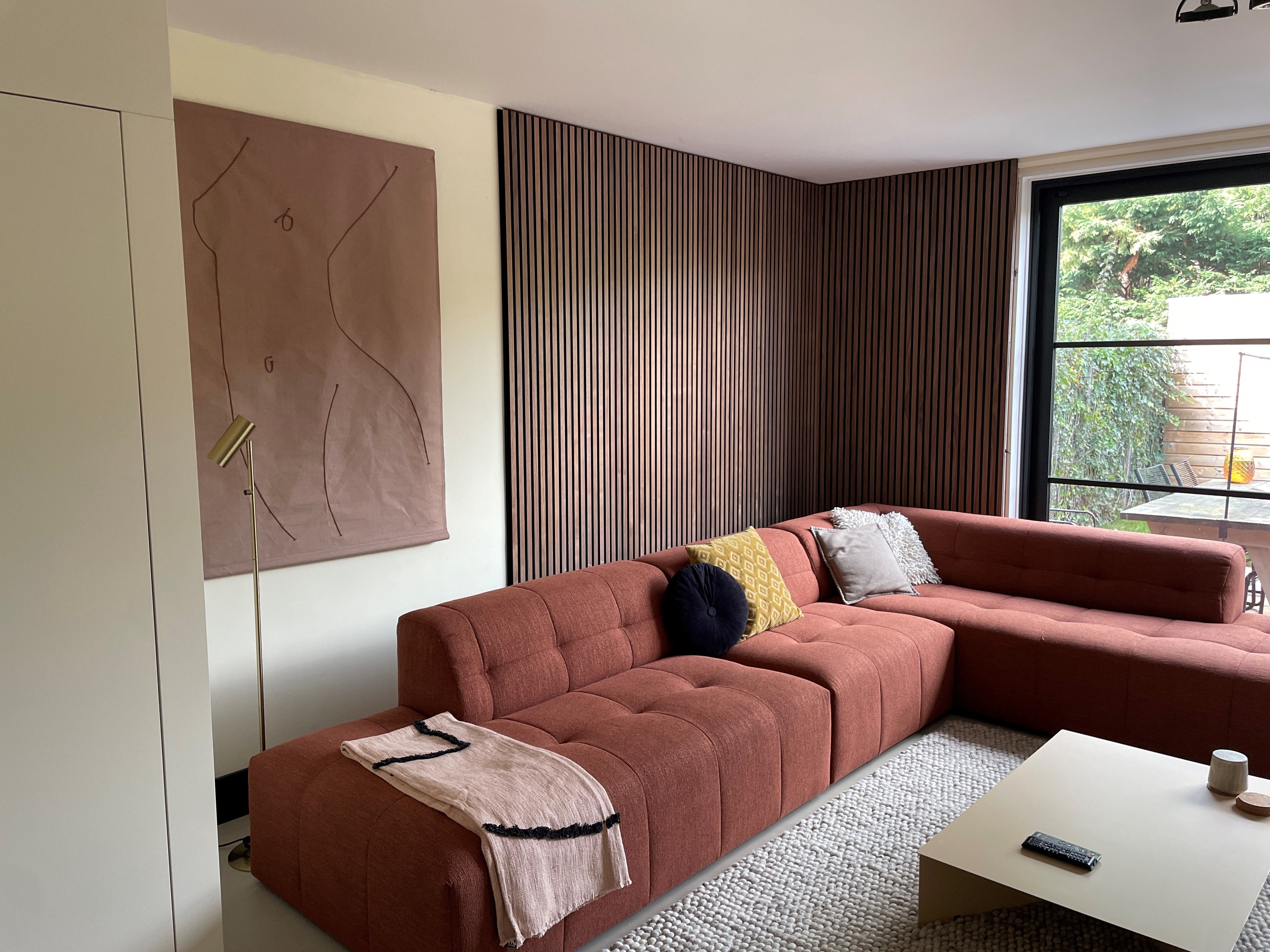 A modern living room with a rust-colored sectional, acoustic wood panels on the wall, abstract art, and a large window overlooking lush greenery.