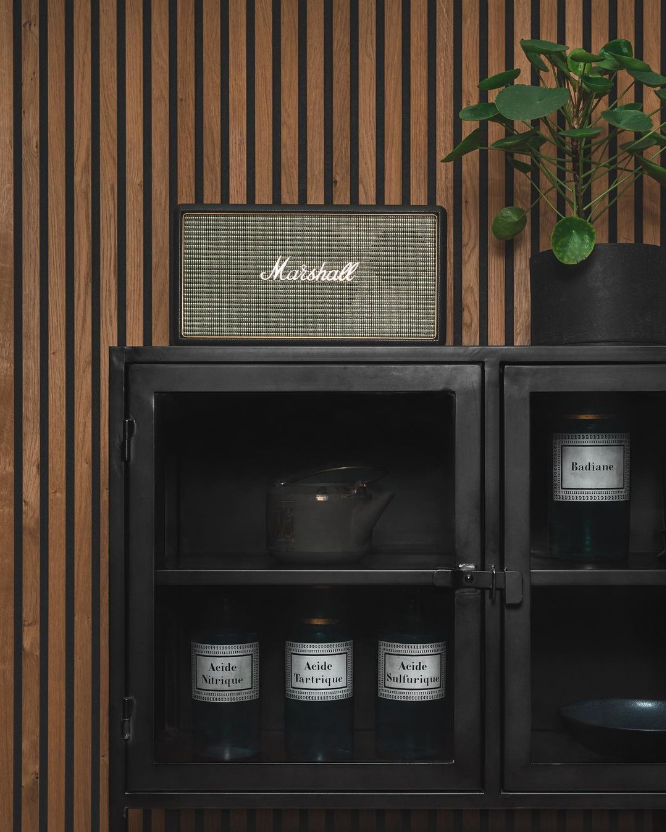 A Marshall Bluetooth speaker sits on a WoodUpp Akupanel acoustic slatwall above a black cabinet holding jars and a potted plant.