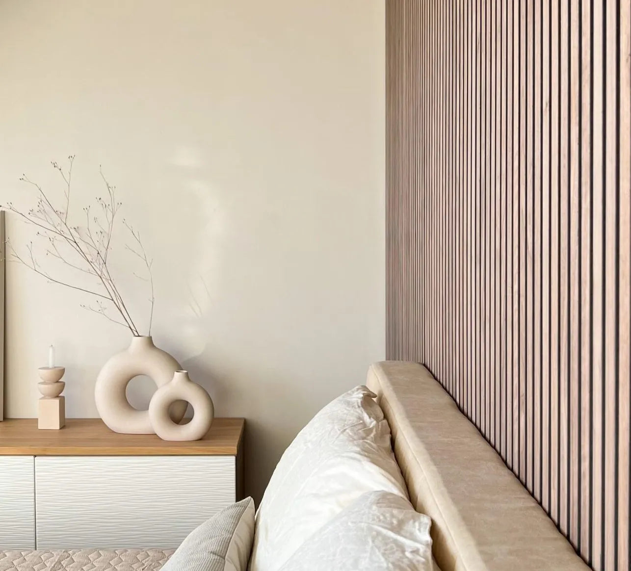 Minimalist bedroom with beige bed, textured Akupanel wooden slat accent wall, and modern vases; neutral tones create a calm, Nordic-inspired atmosphere.